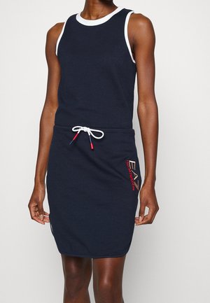 Frau trägt ärmelloses marineblaues Kleid mit weißen Akzenten, Tunnelzug an der Taille und rotem EA7 Emporio Armani Logo am Rock.