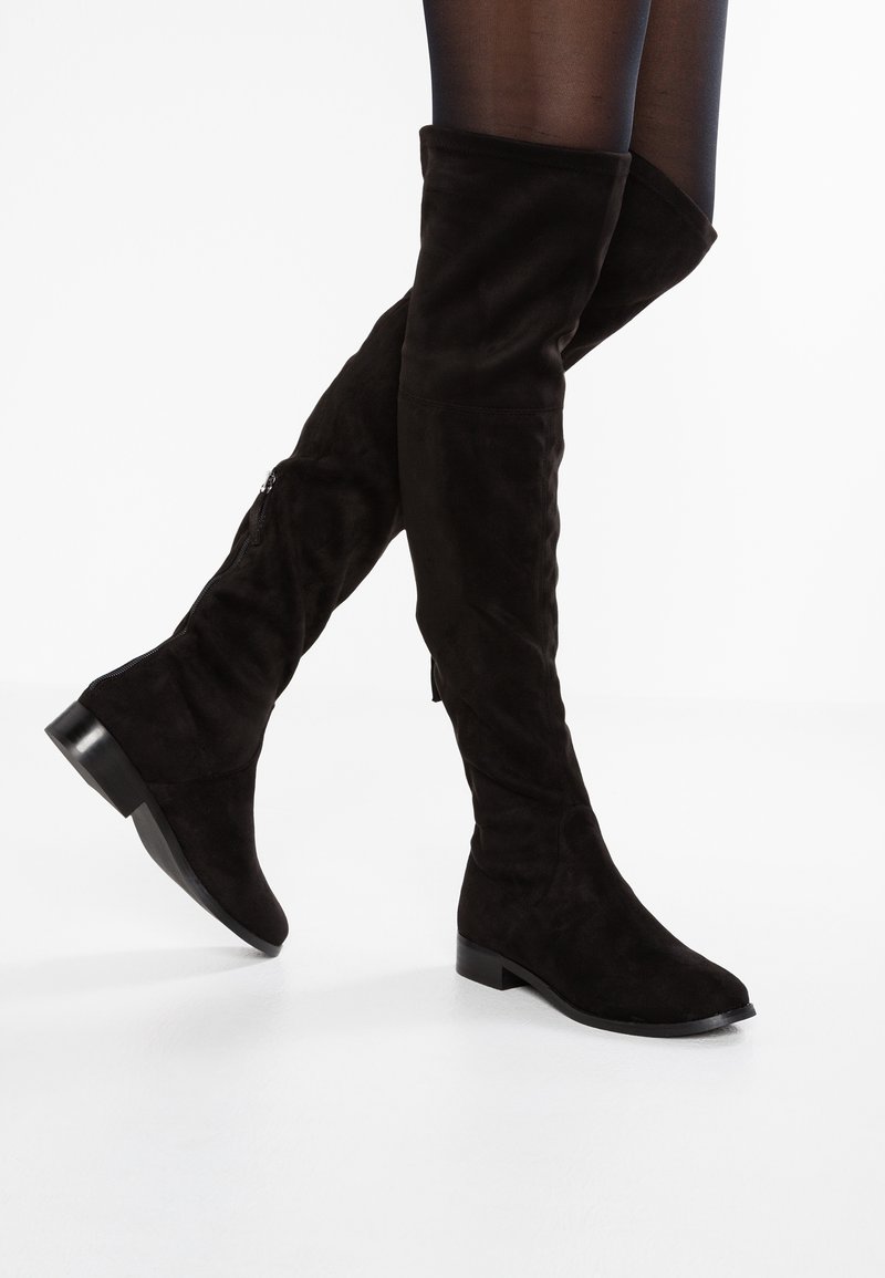 Bottes plates en daim noir mi-cuisses portées avec des collants noirs transparents, présentées avec une jambe croisée derrière l'autre sur fond blanc.