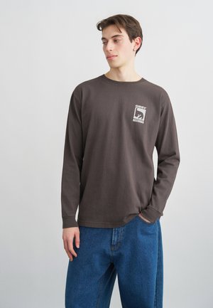 Νεαρός άνδρας με μακρυμάνικο πουκάμισο σκούρου γκρι χρώματος με το λογότυπο "OBEY RECORDS", μπλε τζιν, στέκεται με το ένα χέρι στην τσέπη, κοιτάζοντας προς τα αριστερά.