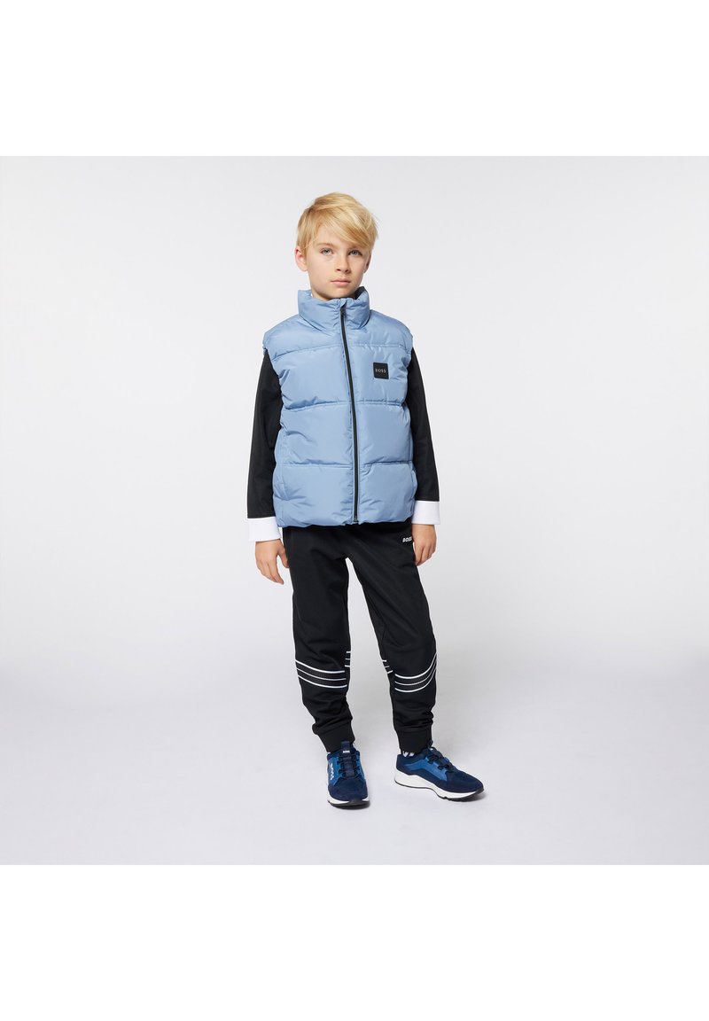 Gilet rembourré bleu clair avec un col haut, des accents noirs et des poches latérales, porté sur une chemise noire à manches longues et un pantalon noir avec des rayures blanches.