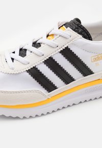 Vit sportskor med svarta ränder, överdel i mocka och mesh, texturerad svart häl, gula detaljer och vit gummisula med räfflor.