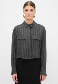 Geknipt zwart shirt met witte verticale strepen. Heeft een kraag, knoopsluiting aan de voorkant en twee borstzakken. Lange mouwen met gestreepte manchetten.