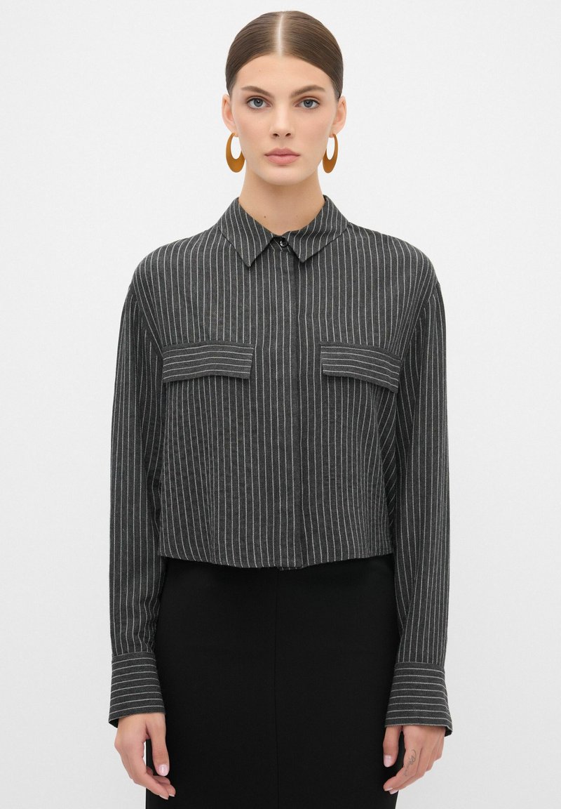 Geknipt zwart shirt met witte verticale strepen. Heeft een kraag, knoopsluiting aan de voorkant en twee borstzakken. Lange mouwen met gestreepte manchetten.