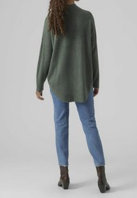 Femme aux cheveux bouclés portant un pull vert surdimensionné, un jean bleu clair et des bottines à talons marron, debout de dos.