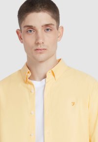 Geel button-up overhemd met een subtiele textuur en een klein geborduurd logo. Draagt over een wit T-shirt, met een traditionele kraagontwerp.