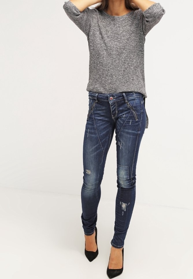 Femme portant un pull gris et un jean skinny bleu foncé déchiré avec des talons hauts noirs, debout devant un fond blanc.