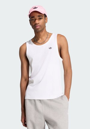 Man draagt een wit mouwloos geribd shirt, lichtgrijze joggingbroek, een roze pet en een zilveren ketting, staand met één hand in de zak.