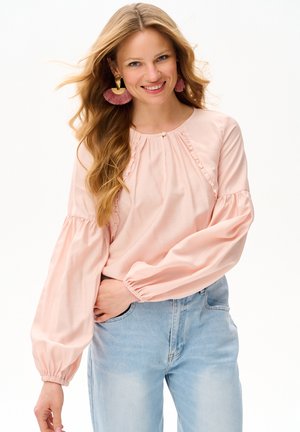 Uśmiechnięta kobieta z długimi blond włosami ma na sobie różową bluzkę z bufkami, złote i różowe kolczyki z frędzlami oraz jasnoniebieskie dżinsy.