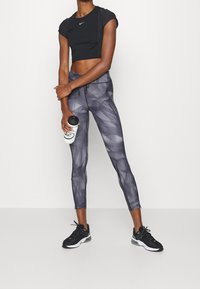 Černý zkrácený top s krátkými rukávy, doplněný šedými legínami s vzorem. Modelka drží lahev vody a má na nohou černé sportovní boty.