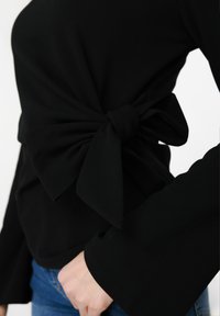 Μαύρο τοπ με μακριά μανίκια και μεγάλο φιόγκο στο πλάι, σε συνδυασμό με τζιν μπλε, εμφανίζοντας μέρος του κορμού και του δεξιού χεριού.