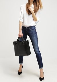 Černá taška přes rameno s horními uchy, vyrobená z hladké kůže. Nositá s bílou košilí s knoflíky, modrými džíny a černými lodičkami se špičatou špičkou.