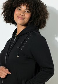 Felpa nera con zip e dettagli allacciati sulle spalle, con occhielli e coulisse argentati. Tessuto morbido e cappuccio accogliente.