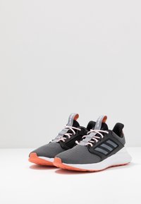 Par de zapatillas Adidas de running grises con suelas melocotón, cordones rosas y rayas laterales negras sobre una superficie blanca y un fondo gris claro.