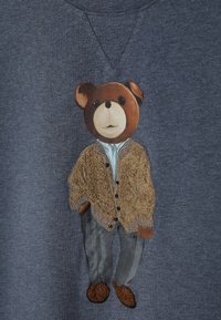 Námořnicky modrá mikina s vyšívaným medvědem oblečeným v hnědém texturovaném kardiganu, světle modré košili, šedých kalhotách a hnědých botách.