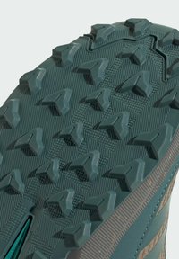 Teal running shoe sole with deep, sharp lugs for traction; textured surface with angular patterns and a reinforced edge.