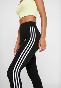 Kvinna iförd svarta Adidas leggings med tre vita ränder och en ljusgul crop top, stående mot en plain ljusgrå bakgrund.