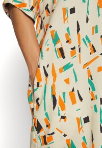 Robe beige avec un motif abstrait comportant des formes orange, verte et noire. Les manches courtes et les poches latérales sont visibles.