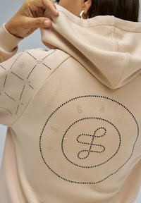 Sudadera con capucha beige con diseño de puntos que presenta un logotipo circular y un patrón geométrico en la manga, acentuada con tachuelas doradas y negras.