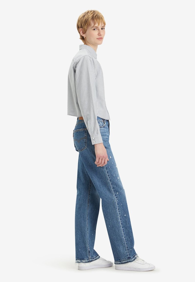 Camicia a righe, corta e con bottoni, di colore azzurro chiaro e bianco; abbinata a jeans larghi e strappati di colore blu e sneakers bianche, vista di lato.