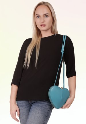 Jonge vrouw met lang blond haar draagt een zwarte top, blauwe spijkerbroek en een blauwe hartvormige crossbodytas op een lichte achtergrond.