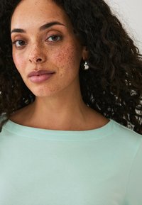 Top verde menta a maniche corte con scollatura ampia, realizzato in un tessuto liscio e leggero. La modella ha i capelli ricci e indossa orecchini a cerchio in argento.