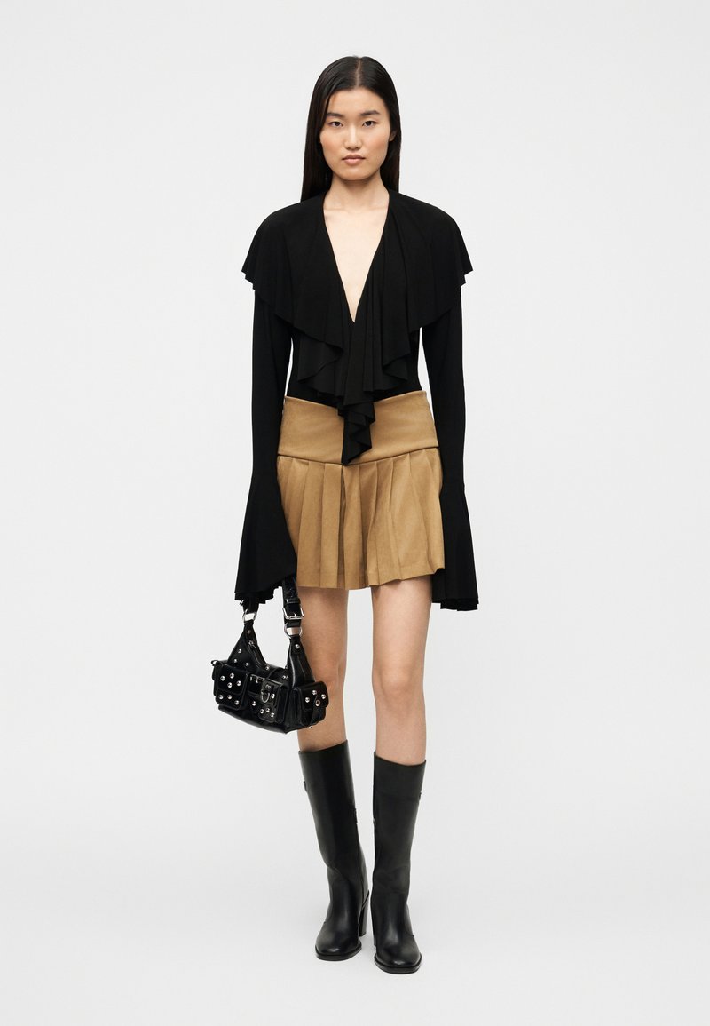 Haut enveloppant noir à volant sur le devant, jupe plissée beige, bottes noires montantes jusqu'aux genoux, et petit sac à main noir avec des clous argentés.