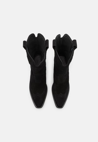 Botines de ante negros hasta el tobillo con punteras afiladas, costuras decorativas y tiradores. Presentan una parte superior texturizada y suelas negras mate.