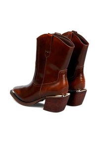 Botas de tobillo de cuero marrón con punta afilada y tacón apilado. Presentan costuras decorativas y un acento metálico en el tacón.