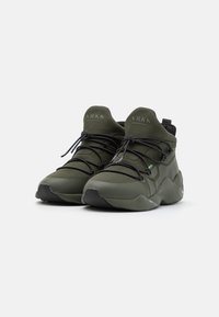 Zapatillas altas verdes con una parte superior de punto texturizado, acentos negros, cordones elásticos y una suela de goma curva, diseñadas para comodidad y soporte.