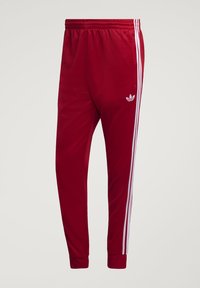 Κόκκινο παντελόνι φόρμας Adidas με λευκές πλαϊνές ρίγες, ελαστική μέση, λάστιχα στους αστραγάλους και το λογότυπο Adidas trefoil στο μηρό.