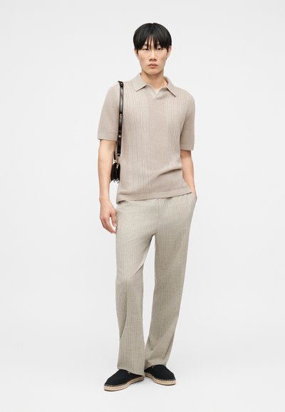 Νεαρός άνδρας που στέκεται, φοράει μπεζ κοντομάνικο πλεκτό πόλο, ανοιχτό μπεζ παντελόνι με φαρδύ πόδι, μαύρα εσπαντρίγια και κρατάει μια σκουρό καφέ τσάντα ώμου.