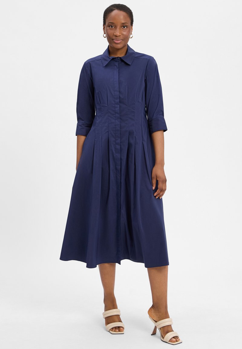 Granatowa bawełniana sukienka z kołnierzykiem, rękawami o długości trzech kwart i plisowaną spódnicą. Posiada zapięcie na guziki z przodu i gładką fakturę.
