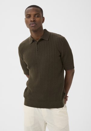 Bărbat purtând un tricou polo texturat, verde măsliniu cu mâneci scurte și pantaloni albi, stând cu mâinile la spate, pe un fundal simplu.