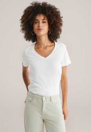 T-shirt blanc à manches courtes en mélange de coton, avec un design ajusté. Porté avec un pantalon de couleur claire et de simples boucles d'oreilles en or.