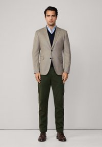 Beige ullblazer över en marinblå tröja, ljusblå skjorta och mörkgröna byxor. Bruna läderskor fulländar outfiten. Enkelt och skräddarsytt design.