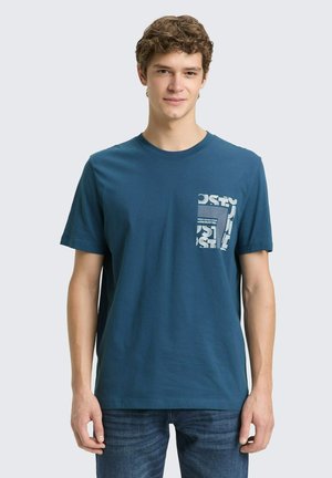 Junger Mann trägt ein blaues T-Shirt mit Rundhalsausschnitt und einem weißen Grafikdruck auf der Brusttasche, kombiniert mit blauen Jeans, steht vor einem schlichten Hintergrund.