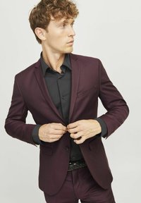 Bordeauxfarbener Anzugjackett mit schmaler Passform, abgesetztem Revers und einer Brusttasche, kombiniert mit einem schwarzen Hemd darunter.