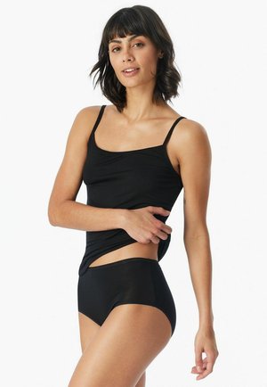 Frau trägt schwarzen Tanktop und Unterwäsche, hebt leicht den Saum des Tanktops, steht vor weißem Hintergrund.