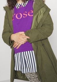 Chaqueta parka verde con borde de piel, llevada sobre una camisa morada con texto "rose", una camisa de rayas con cuello y leggings con patrón abstracto.