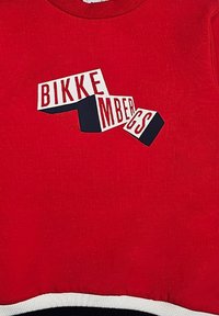 Felpa rossa con lettere grandi bianche e nere che dicono "BIKKENBERGS." Presenta un collo rotondo e un orlo a strisce bianche e nere.