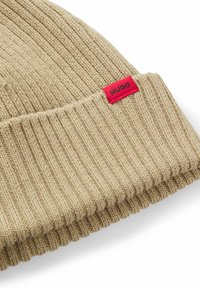 Berretto beige lavorato a maglia con una texture a coste, caratterizzato da un'etichetta rossa con la scritta "HUGO" in rilievo nero. Il design è semplice e aderente.