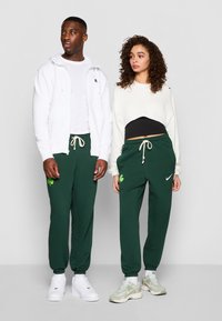Dois modelos usam roupas casuais. O modelo masculino veste um moletom com zíper branco sobre uma t-shirt branca, combinado com calças de jogging verde escuras. A modelo feminina usa um moletom creme cropped sobre uma blusa preta, com calças de jogging verdes que apresentam um desenho de trevo verde. Ambos usam tênis.