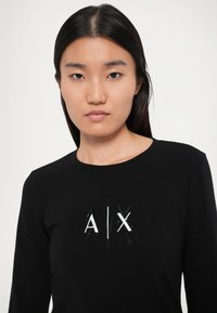 Must mustariga T-särk, millel on keskel valge ja peegelmustast materjalist "A/X" logo. Sile kangas ja ümmargune kaelus.