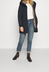 Laivastonsininen parkatakki, jossa on karvavuorittaja huppu, pitkät hihat ja nappikiinnitys; yhdistetty vaaleansinisiin, käännettävien lahkeiden farkkuihin ja tummanruskeisiin nilkkabootseihin.