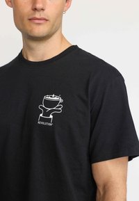 Zwarte katoenen T-shirt met een witte grafiek van een beker en hand op de linkerkant van de borst, met de tekst "REVOLUTION" eronder. Korte mouwen.