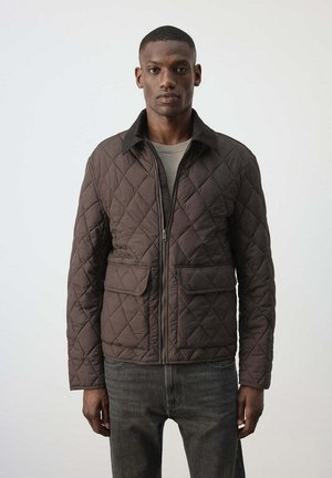 Hombre con chaqueta marrón acolchada con dos bolsillos delanteros con solapa y vaqueros gris oscuro, de pie frente a un fondo claro y liso.