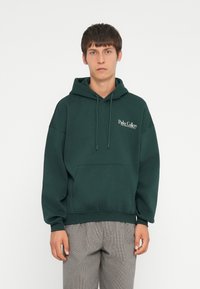 Dunkelgrüne Kapuzenjacke aus Baumwolle mit einer Fronttasche, Kordeln und dem weißen "Parks Gallery"-Logo auf der linken Brust.