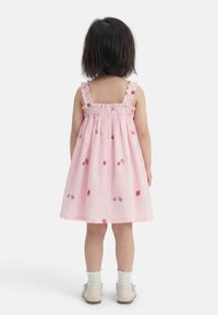 Malček s kratkimi temnimi lasmi, oblečen v roza obleko s potiskom jagod, belimi nogavicami in bež čevlji, stoji obrnjena stran proti belemu ozadju.