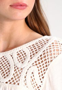 Vit skjorta med en dekorativ virkad halsringning som har en flätad design. Texturen är slät med kontrasterande detaljerad sömnad.