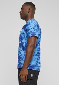 Korst t-skjorte med et blått tie-dye mønster, korte ermer, rund hals, og en logo på forsiden. Modellen har på seg svarte cargo-shorts.
